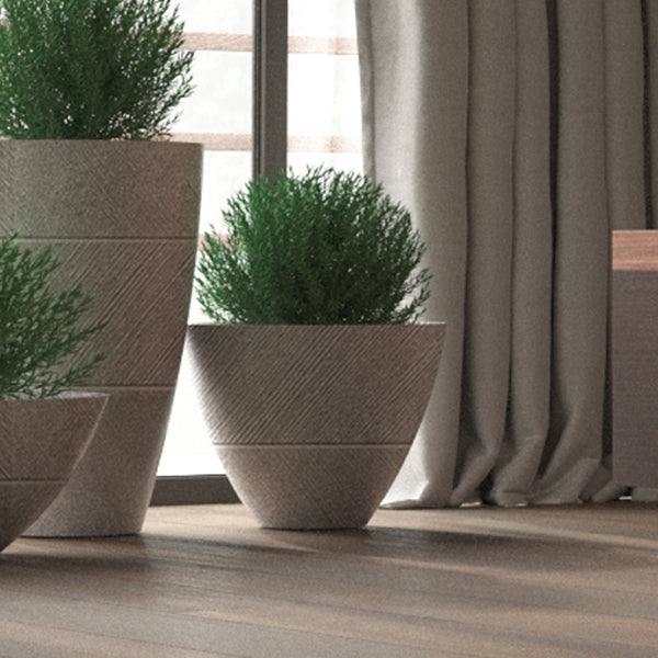 The All Delivery Options Planter - Drizzle - Stone - 18-inch, filled with lush green plants, adds a cozy, natural touch to a space when placed near a window with sheer curtains.