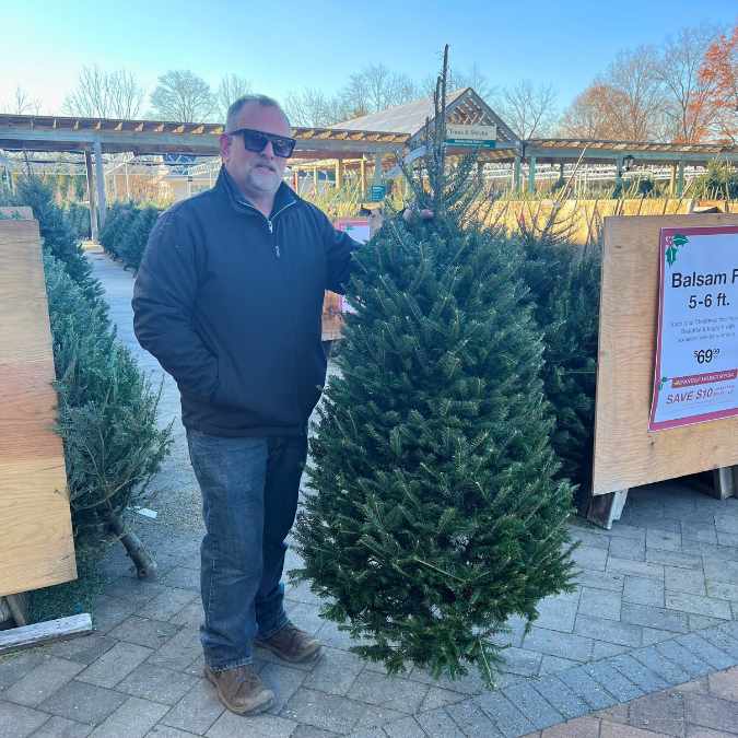 A man in sunglasses and a black jacket stands outdoors beside a Premium Balsam Fir Real Christmas Tree (5-6 ft.), with signs showing prices and more Balsam Fir trees from Available for Pick-up & Delivery in the background.